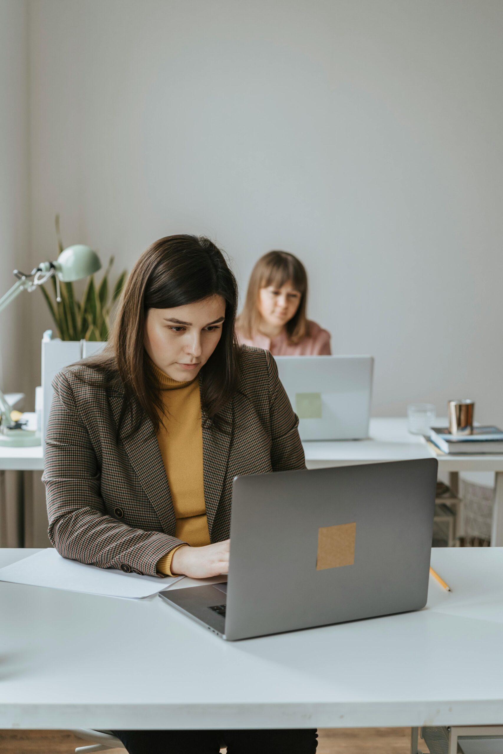 Two women working intently at laptops in a modern office setting.